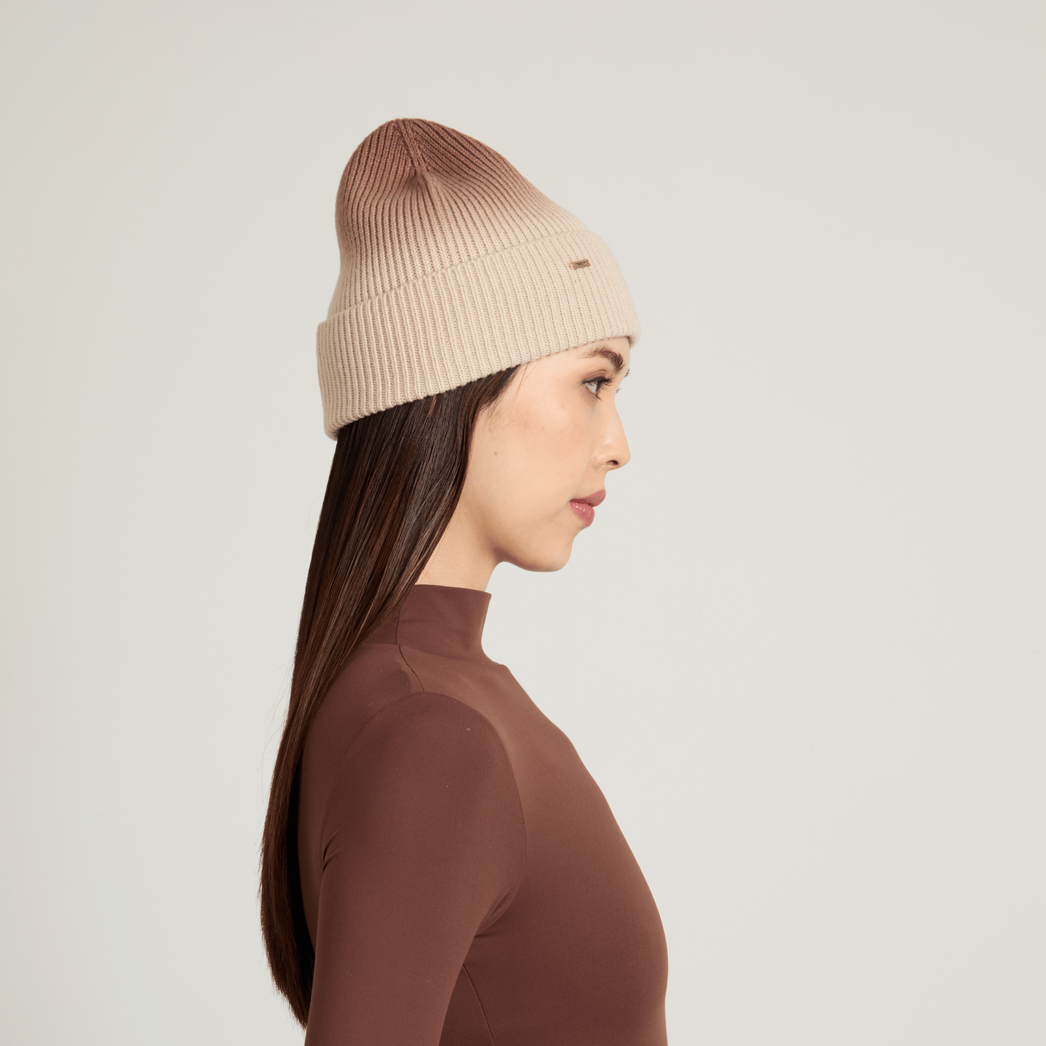 Side view of woman wearing a beige knit ombre beanie against a light gray background