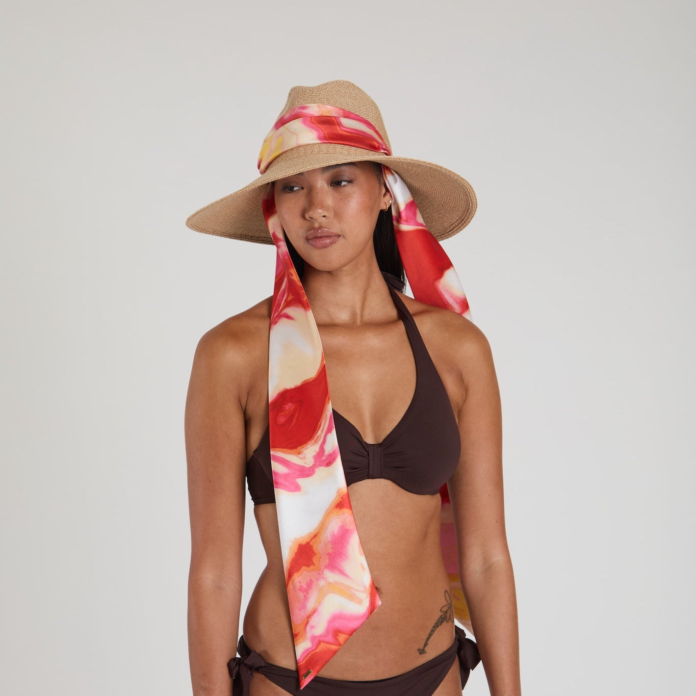 Woman wearing a brown bikini and straw hat with a colorful scarf on a plain background