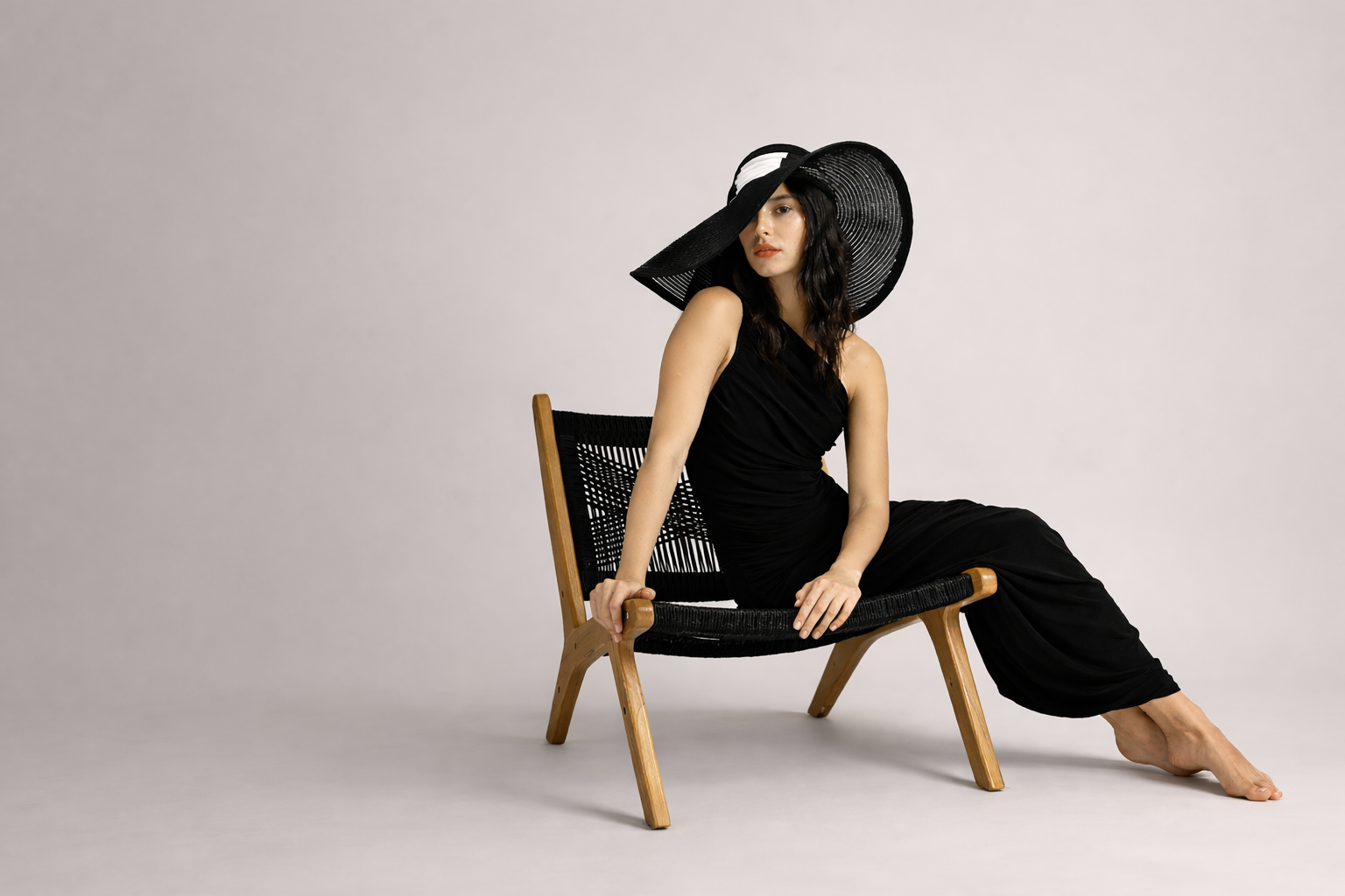 Woman in a black outfit and hat sitting on a wooden chair against a plain background