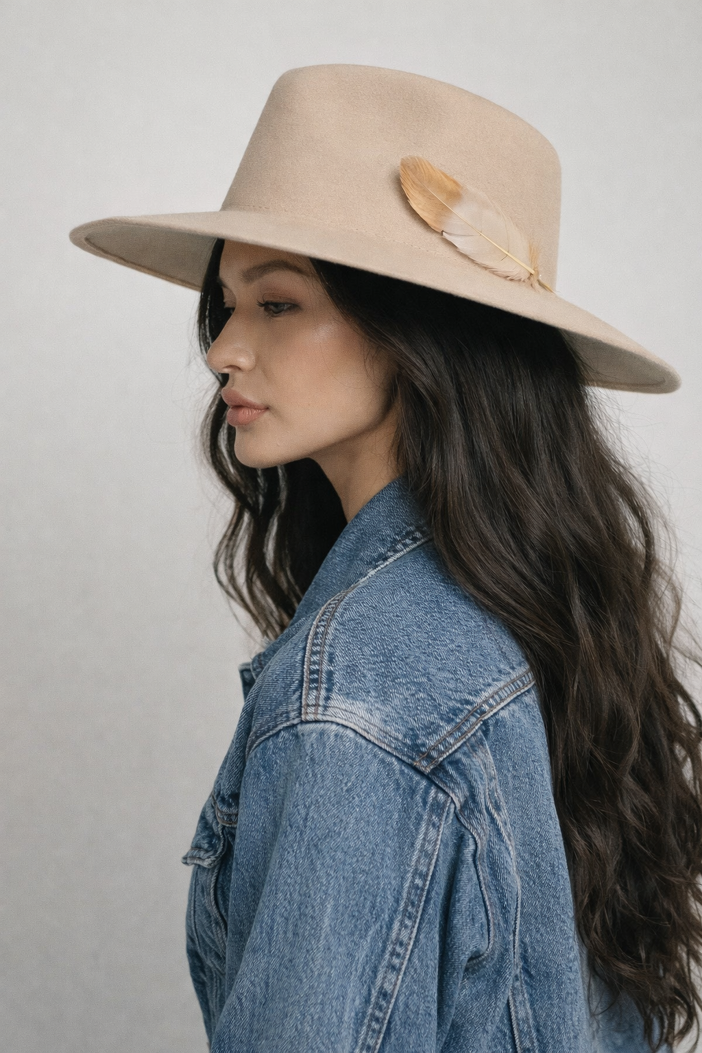 Woman wearing a beige hat with a feather and a denim jacket on a plain background