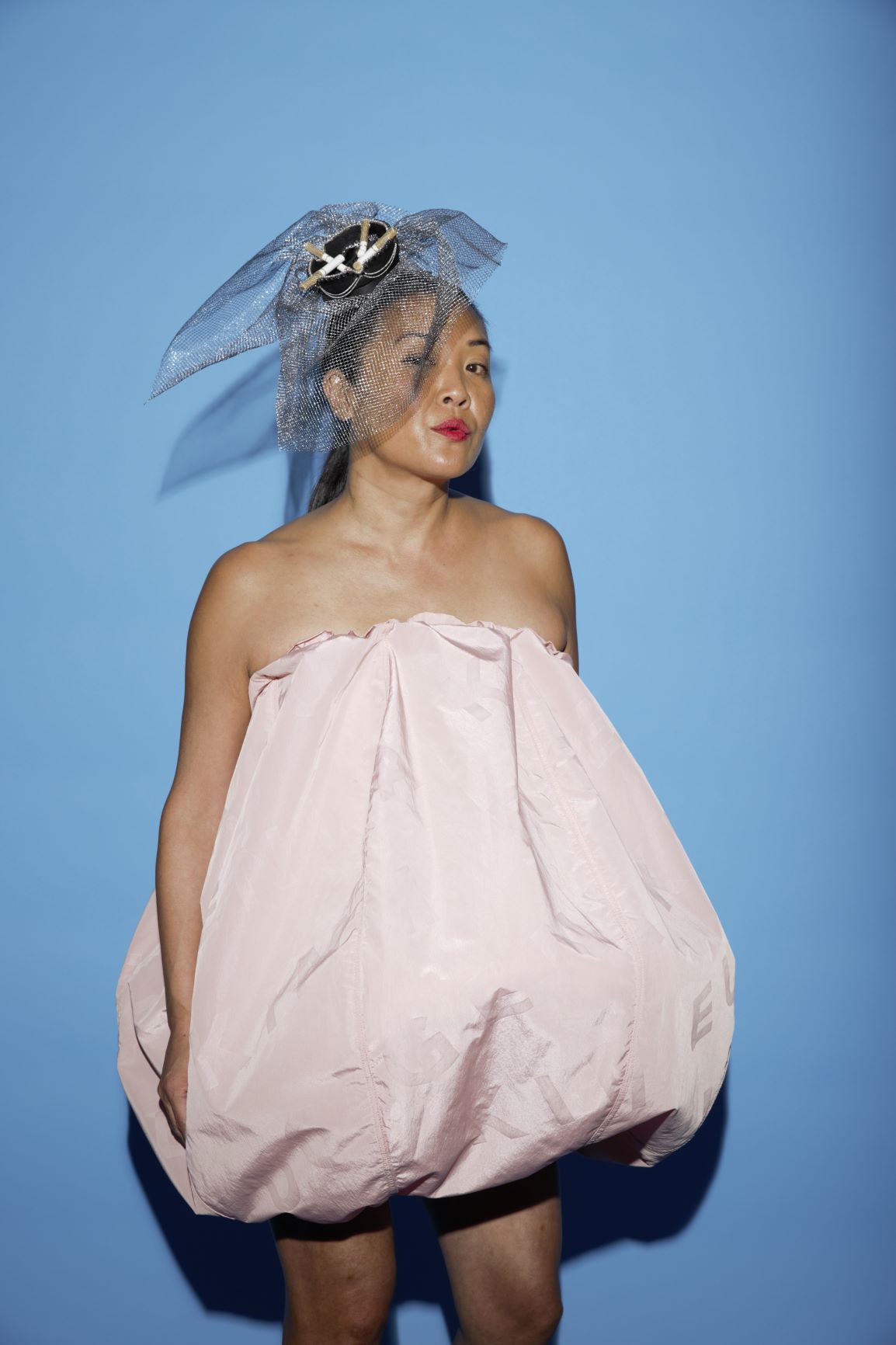 Milliner Eugenia Kim in a pink voluminous bubble dress and a signature headpiece with netting, posing for a fashion event photo.