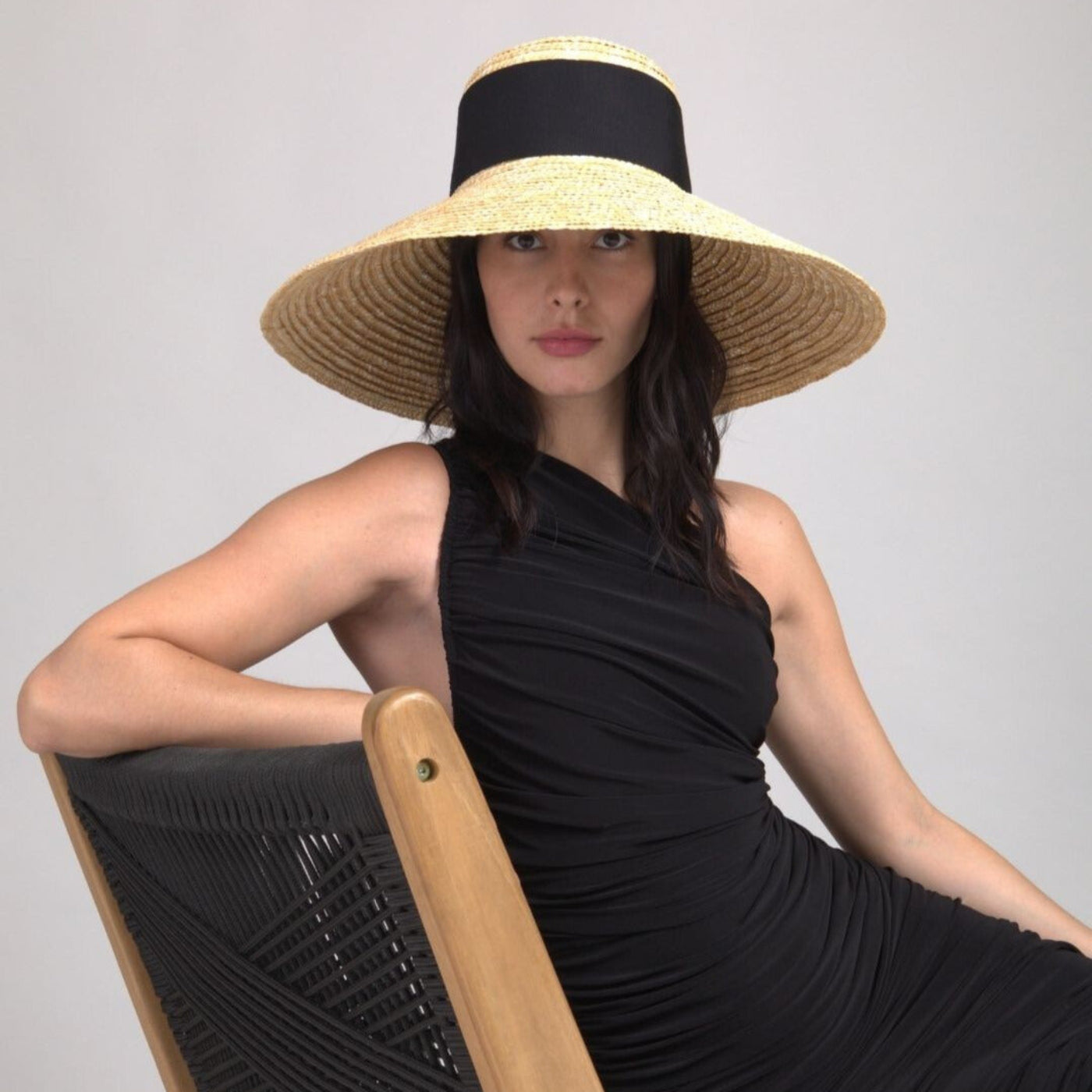 Woman wearing a black dress and wide-brimmed straw hat sitting on a chair against a gray background